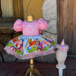 May include: A pink dress with a heart-shaped applique on the bodice and a colorful cartoon print skirt. The dress has puffy sleeves and a tulle hem. A matching party hat with a tulle trim sits on a mannequin head.