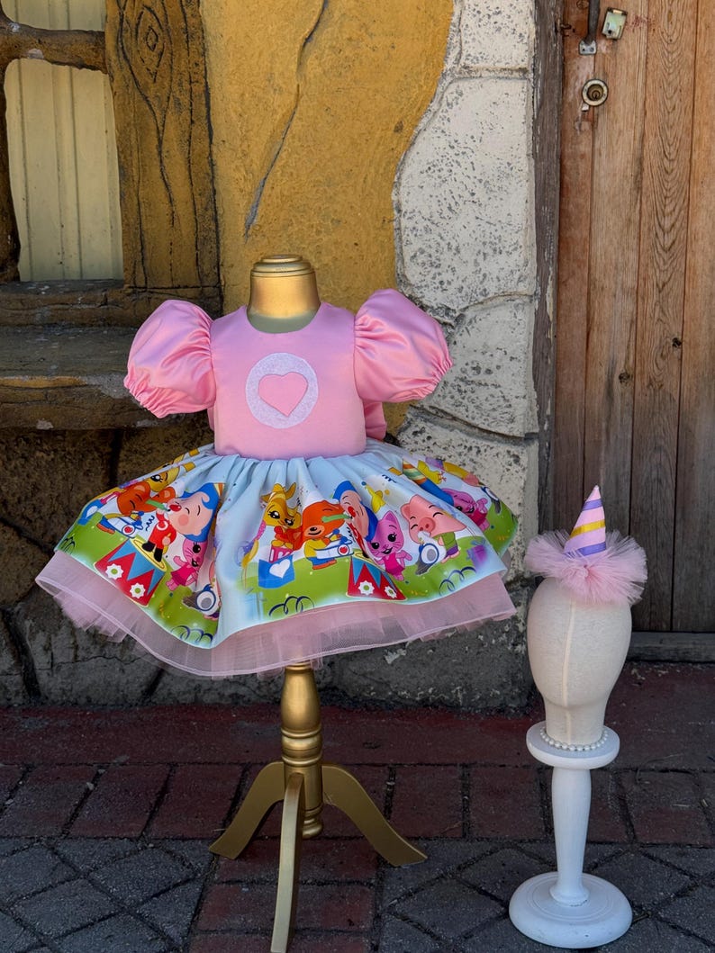 May include: A pink dress with a heart-shaped applique on the bodice and a colorful cartoon print skirt. The dress has puffy sleeves and a tulle hem. A matching party hat with a tulle trim sits on a mannequin head.