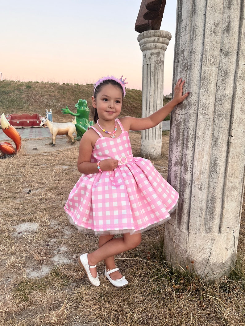May include: A young girl wearing a pink and white gingham dress with a tulle skirt, a pink headband with a crown, and white shoes. She is standing next to a stone pillar.