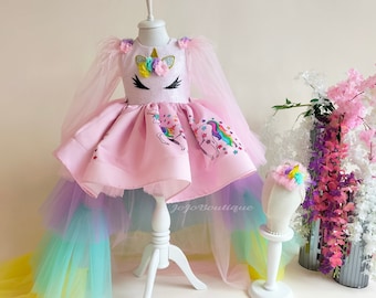 May include: A pink unicorn dress with a tulle skirt in rainbow colors. The dress has a white bodice with a unicorn face and flowers. The dress is on a white stand. Jojo Boutique is printed on the dress.