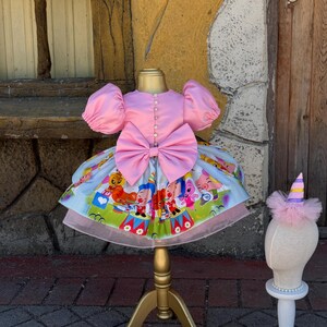 May include: A pink dress with a large bow and puff sleeves is displayed on a gold mannequin. The dress features a colorful circus-themed skirt. A small party hat with a pink tulle base sits on a separate mannequin head.