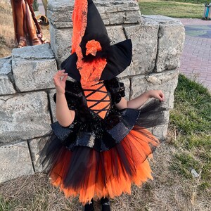 May include: A child dressed as a witch for Halloween. The costume includes a black and orange tulle skirt, a black sequined bodice, and a black witch hat with orange lace trim.