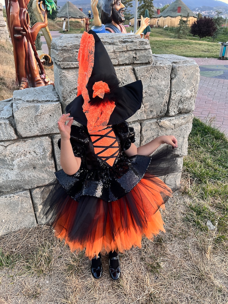 May include: A child dressed as a witch for Halloween. The costume includes a black and orange tulle skirt, a black sequined bodice, and a black witch hat with orange lace trim.