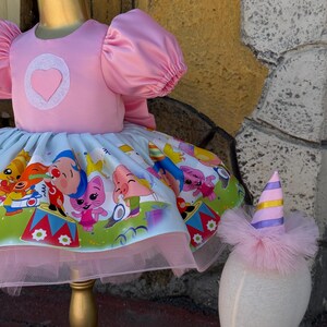 May include: A pink dress with a heart-shaped applique and puffy sleeves. The skirt features a colorful cartoon print and a pink tulle underskirt. A matching party hat with pink feathers and a striped top completes the ensemble. The dress is displayed on a gold mannequin.