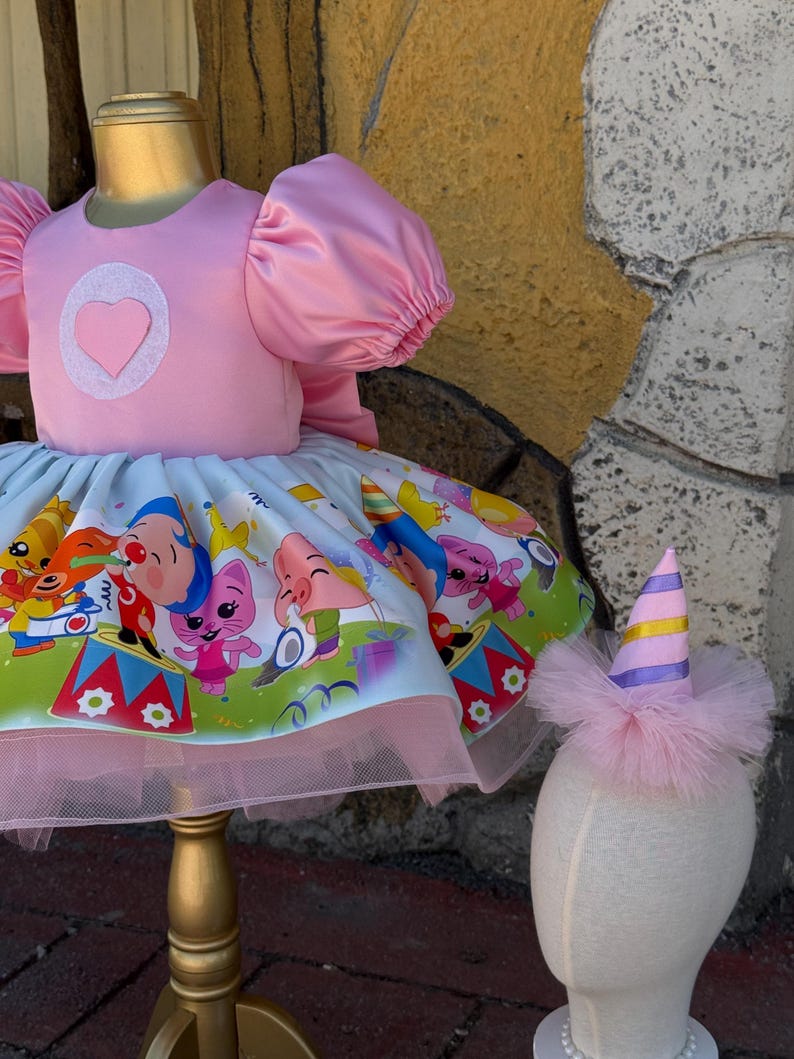 May include: A pink dress with a heart-shaped applique and puffy sleeves. The skirt features a colorful cartoon print and a pink tulle underskirt. A matching party hat with pink feathers and a striped top completes the ensemble. The dress is displayed on a gold mannequin.