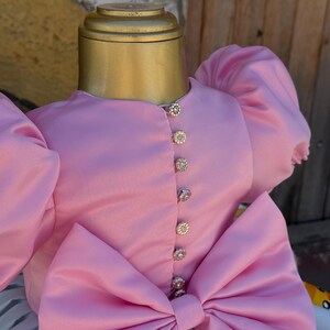 May include: A pink satin dress with large puff sleeves and a large bow. The dress features decorative jeweled buttons down the front. The dress is displayed on a mannequin.