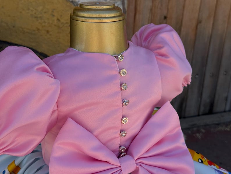 May include: A pink satin dress with large puff sleeves and a large bow. The dress features decorative jeweled buttons down the front. The dress is displayed on a mannequin.