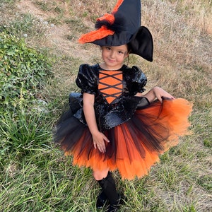 May include: A child dressed as a witch for Halloween. The costume includes a black and orange tulle skirt, a black sequined top with orange accents, and a black witch hat with orange lace trim.