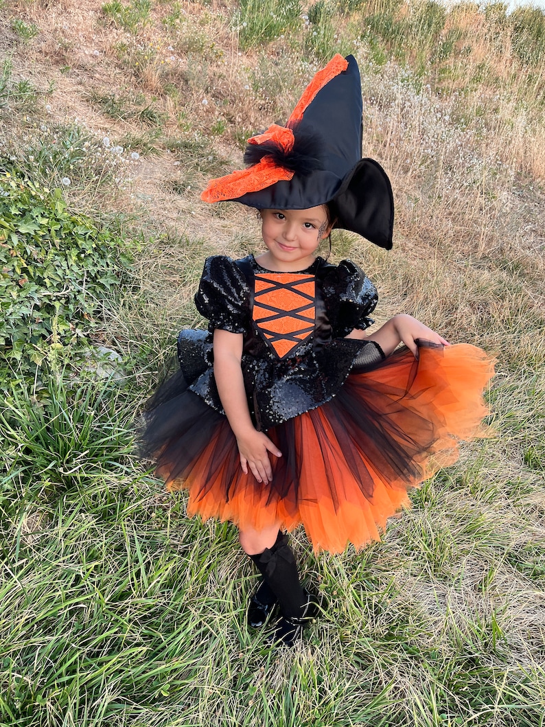 May include: A child dressed as a witch for Halloween. The costume includes a black and orange tulle skirt, a black sequined top with orange accents, and a black witch hat with orange lace trim.