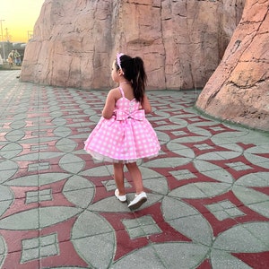 May include: A young girl wearing a pink and white gingham dress with a white bow on the back. She is walking on a patterned sidewalk.
