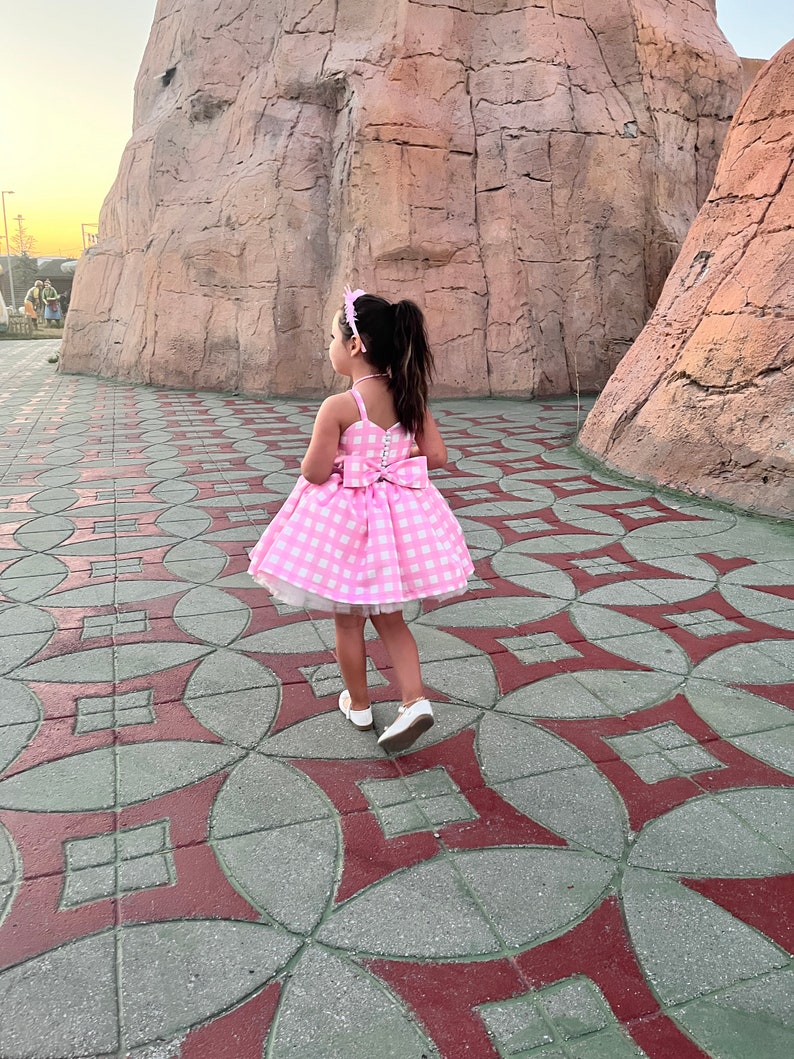 May include: A young girl wearing a pink and white gingham dress with a white bow on the back. She is walking on a patterned sidewalk.