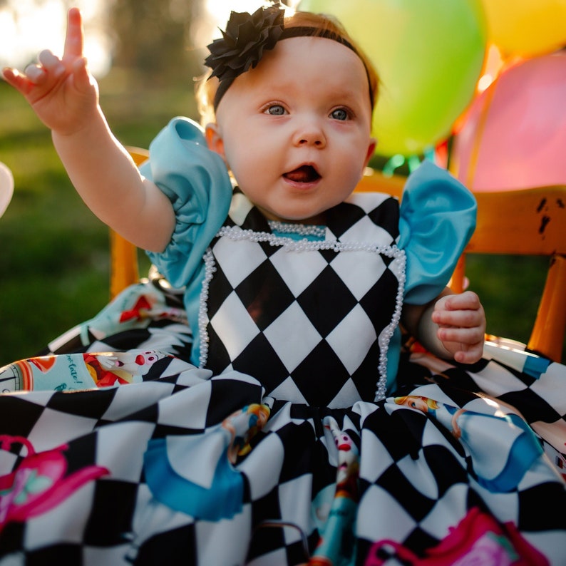May include: A baby girl wearing a blue and white checkered dress with a black and white diamond pattern. She is wearing a black headband with a flower on top. The dress has a blue bodice with white lace trim. The baby is sitting on a wooden chair with a pink and white balloon in the background.