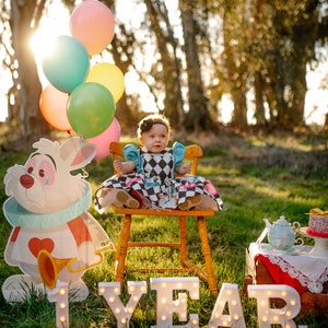 May include: A baby girl wearing a blue and white checkered dress sits on a wooden chair in a grassy field. She is surrounded by colorful balloons and a white rabbit cutout. The words "1 YEAR" are spelled out in white letters with lights on the grass.