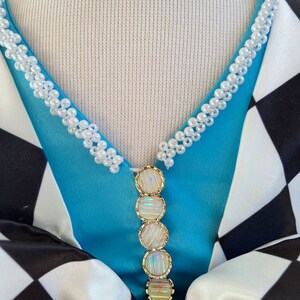May include: A blue and white checkered fabric with a white pearl trim around the neckline. A gold and iridescent decorative piece hangs from the neckline.