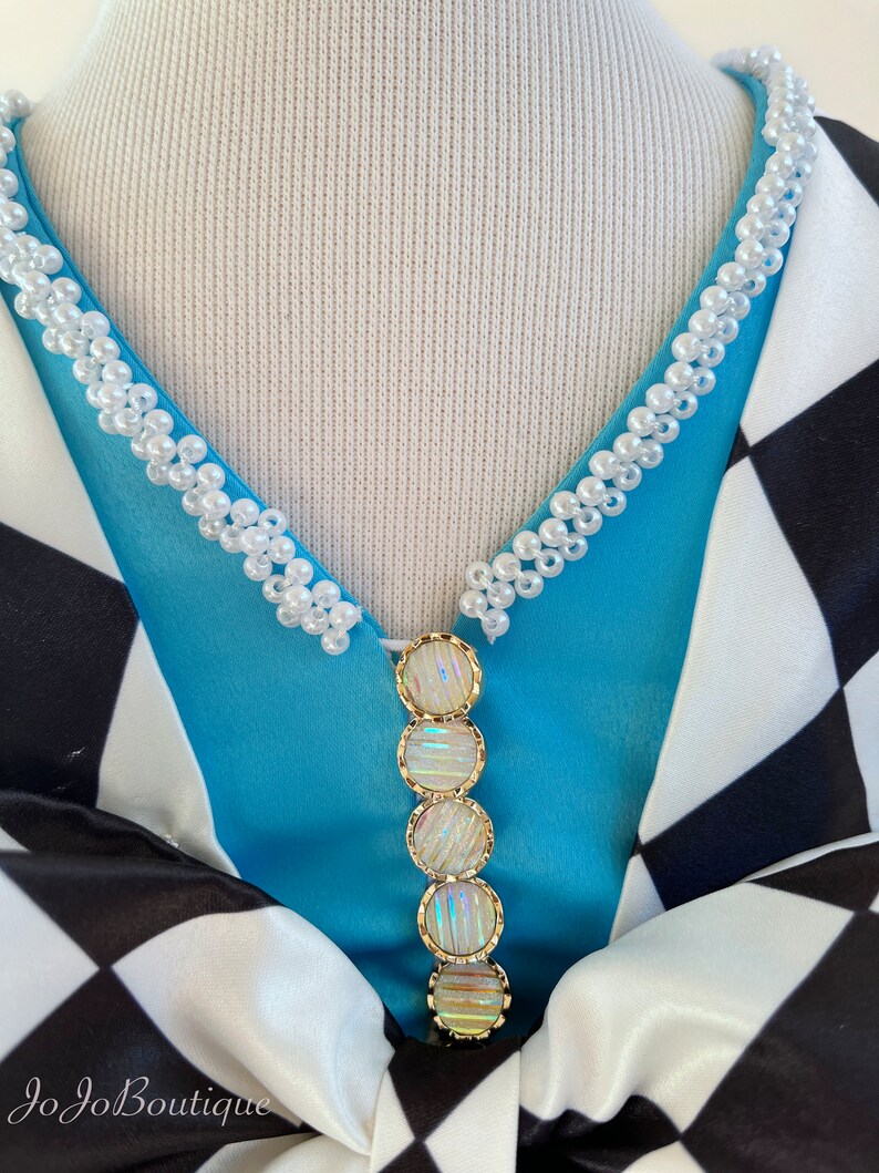 May include: A blue and white checkered fabric with a white pearl trim around the neckline. A gold and iridescent decorative piece hangs from the neckline.