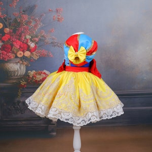 Op de afbeelding: Een gele en witte jurk met een rood, blauw en gouden lijfje. De jurk heeft een kantversiering en een grote strik op de rug.