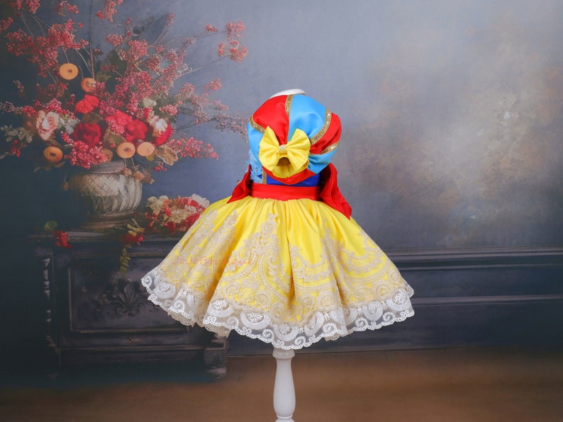 Op de afbeelding: Een gele en witte jurk met een rood, blauw en gouden lijfje. De jurk heeft een kantversiering en een grote strik op de rug.