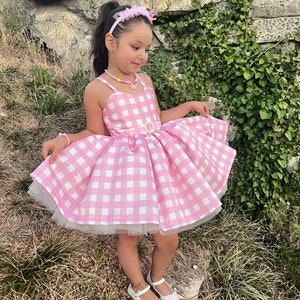 May include: A young girl wearing a pink and white gingham dress with a white tulle underskirt. She is wearing a pink headband with a flower and a colorful beaded necklace. She is standing on a rock with green grass and vines in the background.