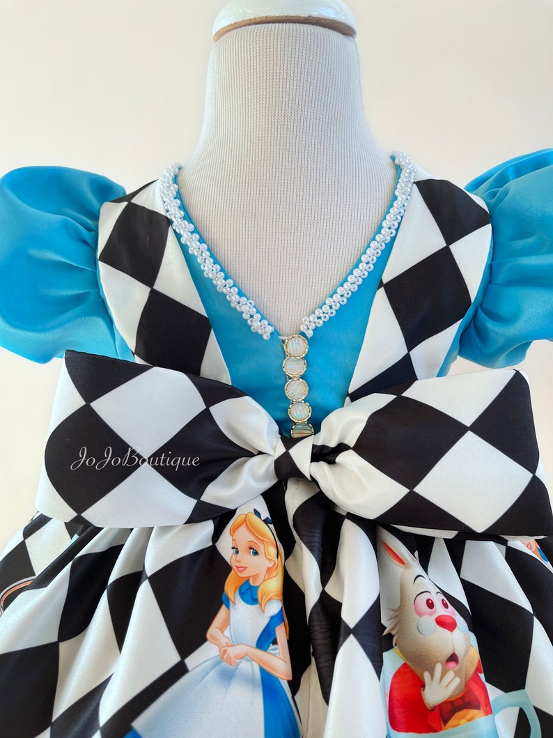 May include: A blue and white checkered dress with a blue bodice and a white pearl necklace. The dress has a large bow in the back and features Alice in Wonderland characters.