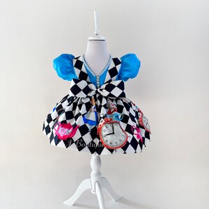 May include: A white and black checkered dress with a blue bodice and a white pearl trim. The dress features a bow at the waist and a print of Alice in Wonderland characters and objects, including a teapot, a pocket watch, and a playing card.
