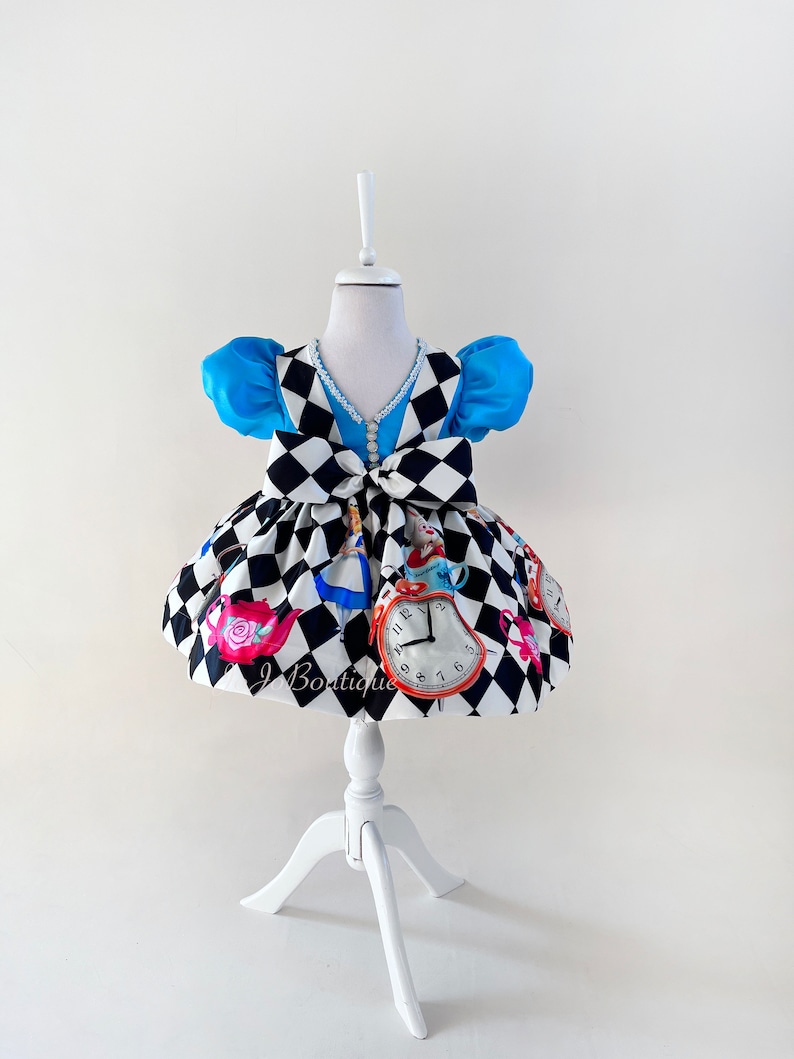 May include: A white and black checkered dress with a blue bodice and a white pearl trim. The dress features a bow at the waist and a print of Alice in Wonderland characters and objects, including a teapot, a pocket watch, and a playing card.