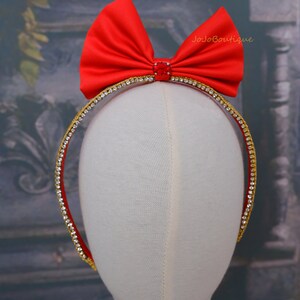 Op de afbeelding: Een rood satijnen strik hoofdband met een gouden en strass rand. Het hoofdband is op een witte mannequin hoofd.