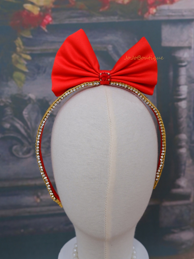 Op de afbeelding: Een rood satijnen strik hoofdband met een gouden en strass rand. Het hoofdband is op een witte mannequin hoofd.