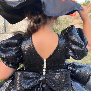 May include: Black sequined dress with a large bow in the back. The dress has puff sleeves and a V-neckline. The dress is adorned with pearl-like buttons down the back.