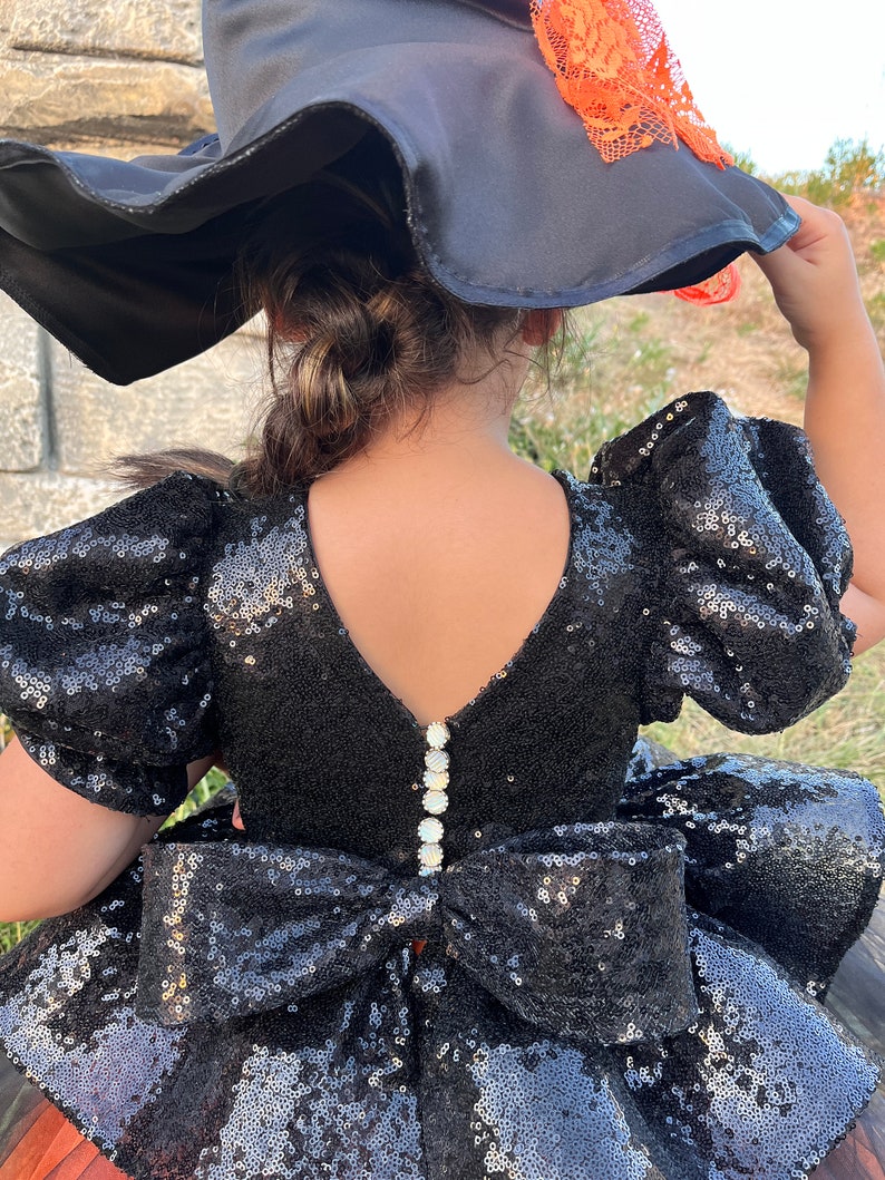 May include: Black sequined dress with a large bow in the back. The dress has puff sleeves and a V-neckline. The dress is adorned with pearl-like buttons down the back.