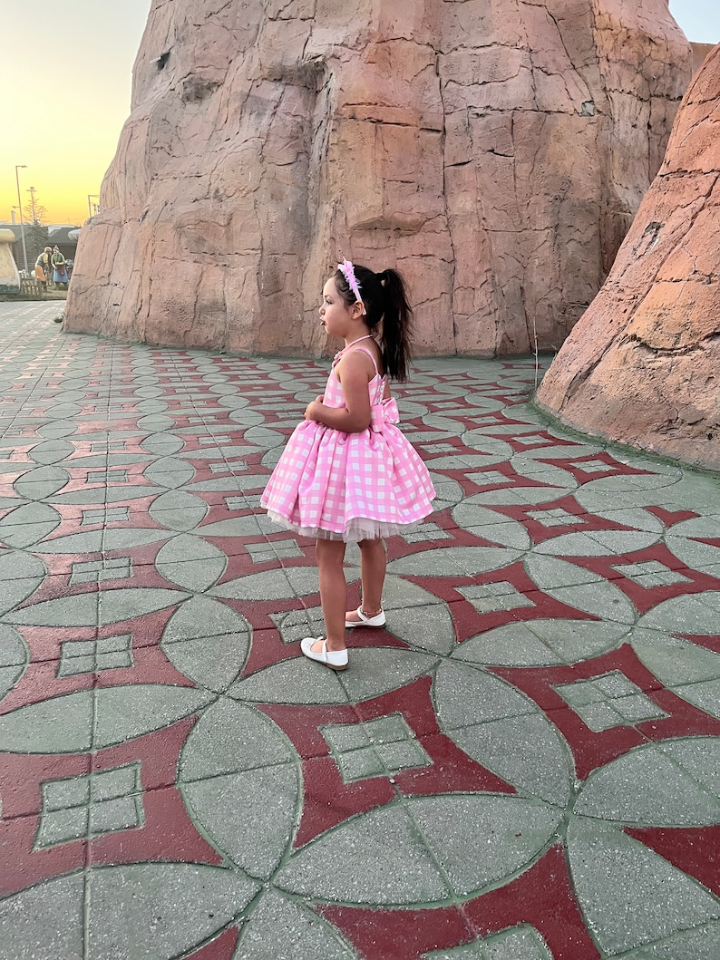 May include: A young girl wearing a pink and white gingham dress with a white tulle underskirt. She is standing on a patterned tile floor.