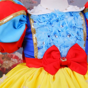 Op de afbeelding: Een gele, blauwe, rode en witte jurk met een grote rode strik en een blauw en wit gerimpeld lijfje. De jurk heeft een witte kraag met parel accenten.