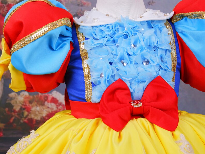 Op de afbeelding: Een gele, blauwe, rode en witte jurk met een grote rode strik en een blauw en wit gerimpeld lijfje. De jurk heeft een witte kraag met parel accenten.