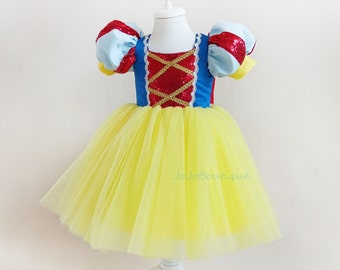 Puede incluir: Un vestido de tul amarillo con un corpiño azul y rojo con un diseño de lentejuelas rojas y ribetes dorados. El vestido tiene mangas abullonadas con ribetes blancos. El vestido está en un maniquí blanco.