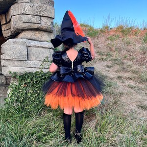 May include: A child wearing a black and orange witch costume with a black hat and a tulle skirt. The costume is decorated with sequins and the hat has a red and orange feather.