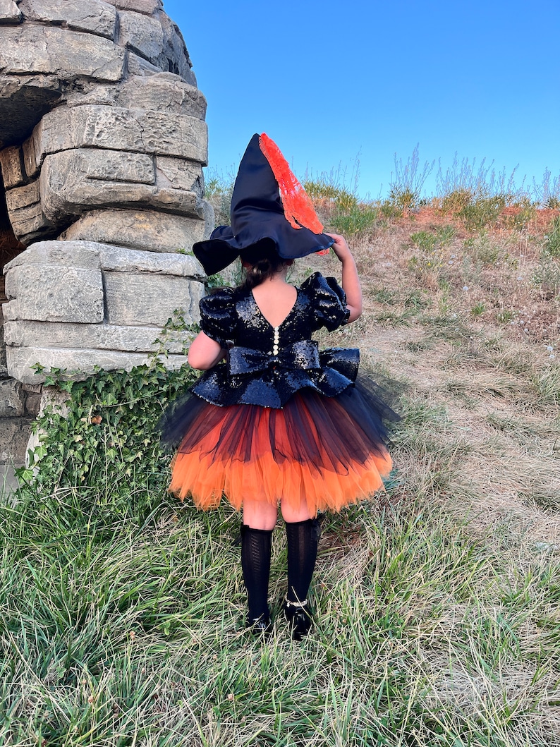May include: A child wearing a black and orange witch costume with a black hat and a tulle skirt. The costume is decorated with sequins and the hat has a red and orange feather.