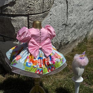 May include: A pink satin dress with a large bow and puff sleeves, displayed on a gold stand. The skirt features a colorful circus-themed print. A matching party hat with a pink trim sits on a white stand.