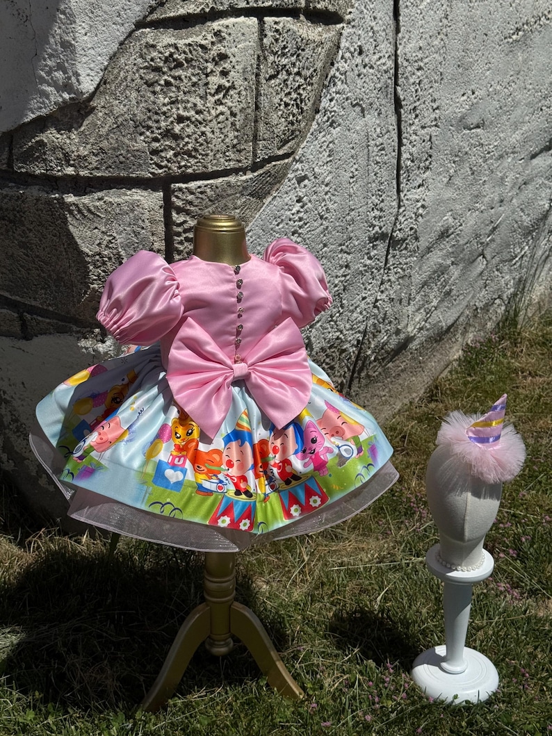May include: A pink satin dress with a large bow and puff sleeves, displayed on a gold stand. The skirt features a colorful circus-themed print. A matching party hat with a pink trim sits on a white stand.