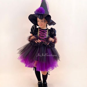 May include: A young girl dressed as a witch for Halloween. She is wearing a black and purple tulle tutu, a black sequined top with a purple ribbon, and a black witch hat with a purple bow. She is also wearing black tights and black shoes.