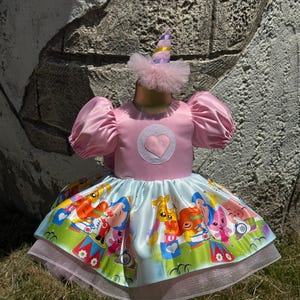 May include: A pink satin dress with a heart applique and puff sleeves. The skirt features a colorful cartoon print and a layered tulle hem. A matching party hat with a tulle trim completes the outfit. This dress is perfect for a birthday party.