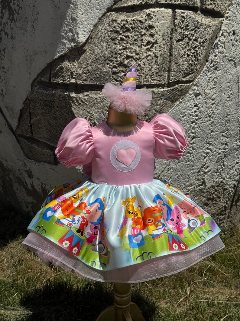 May include: A pink satin dress with a heart applique and puff sleeves. The skirt features a colorful cartoon print and a layered tulle hem. A matching party hat with a tulle trim completes the outfit. This dress is perfect for a birthday party.