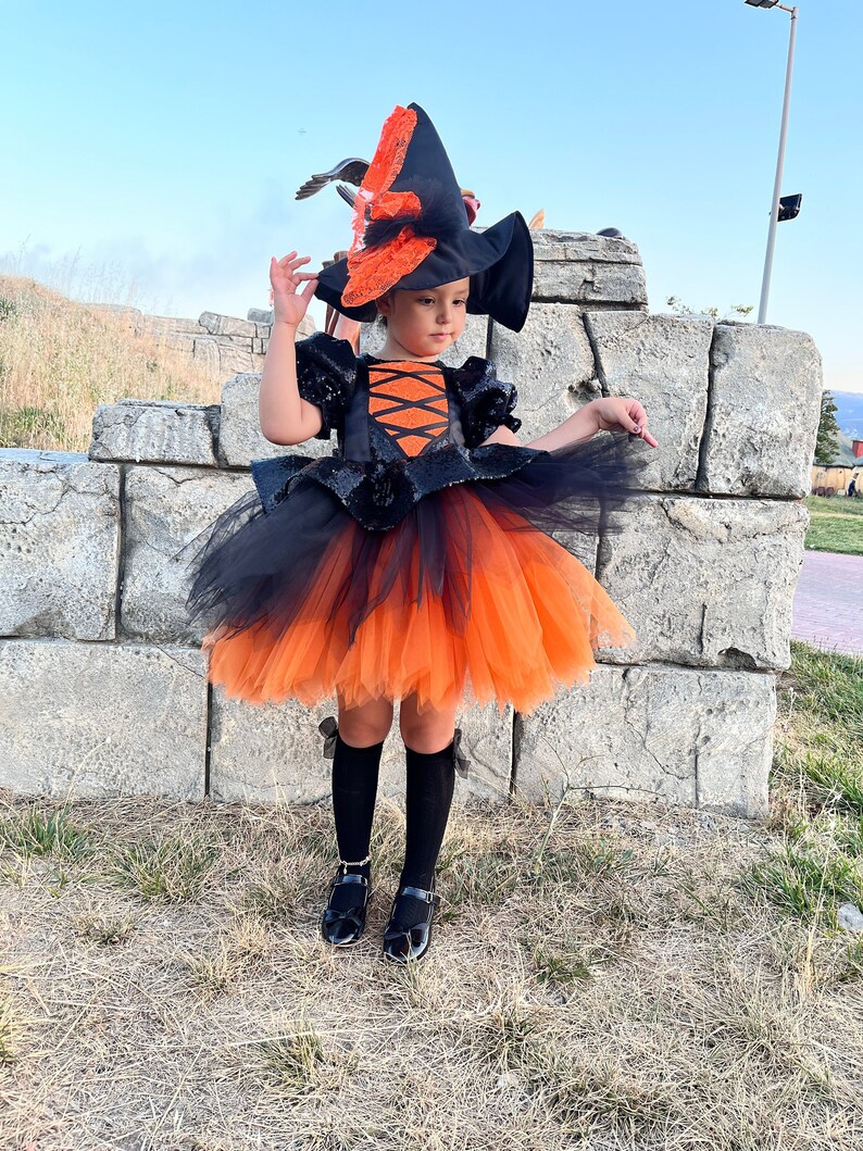 May include: A young girl dressed as a witch for Halloween. She is wearing a black and orange tulle dress with a black sequined bodice and a black witch hat with orange accents. She is also wearing black tights and black shoes.