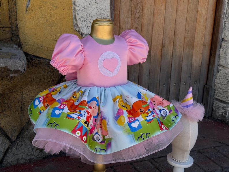May include: Pink toddler dress with puff sleeves and a heart applique. The skirt features a colorful print of cartoon characters. A matching party hat with a pink tulle base sits nearby. The dress is displayed on a mannequin.