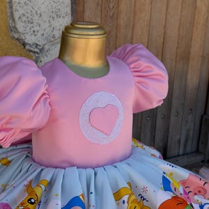 May include: A pink dress with puffy sleeves and a heart-shaped design on the bodice. The skirt features a colorful print with cartoon characters. The dress is displayed on a mannequin, suggesting it's children's apparel.