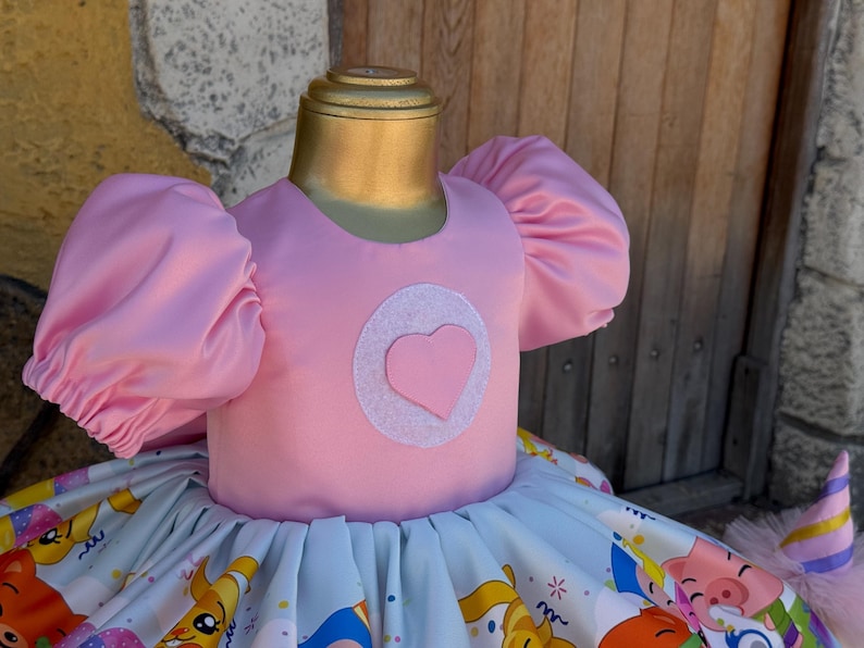 May include: A pink dress with puffy sleeves and a heart-shaped design on the bodice. The skirt features a colorful print with cartoon characters. The dress is displayed on a mannequin, suggesting it's children's apparel.