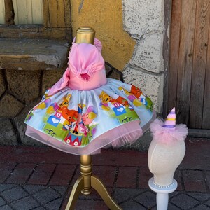 May include: A pink and white dress with a colorful cartoon print, displayed on a gold dress form. The dress has a tulle skirt and a matching pink hat. A smaller mannequin head displays a matching party hat.