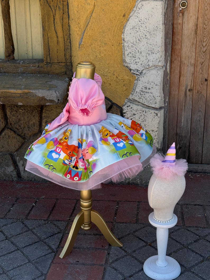 May include: A pink and white dress with a colorful cartoon print, displayed on a gold dress form. The dress has a tulle skirt and a matching pink hat. A smaller mannequin head displays a matching party hat.
