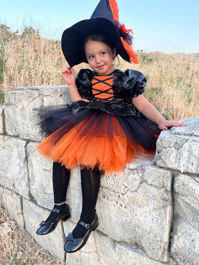 May include: A young girl dressed as a witch for Halloween. She is wearing a black and orange tulle dress with a black sequined bodice, a black witch hat with orange trim, and black shoes. She is sitting on a stone wall.
