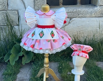 Op de afbeelding: Een roze en witte jurk met aardbeienthema en een bijpassend mutsje. De jurk heeft een rok met aardbeiprint, een wit lijfje en rode accenten. Het mutsje heeft een aardbeienontwerp en rode rand. De jurk wordt getoond op een gouden mannequin.