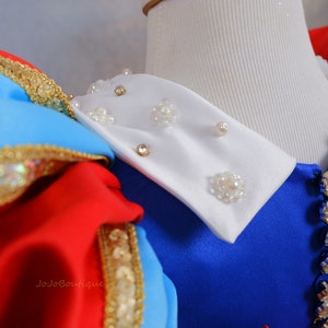 Op de afbeelding: Een witte kraag met parel- en strassversiering, bevestigd aan een blauwe jurk met een rood en blauw lijfje. De jurk heeft gouden versieringen.