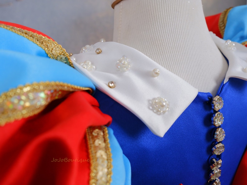 Op de afbeelding: Een witte kraag met parel- en strassversiering, bevestigd aan een blauwe jurk met een rood en blauw lijfje. De jurk heeft gouden versieringen.